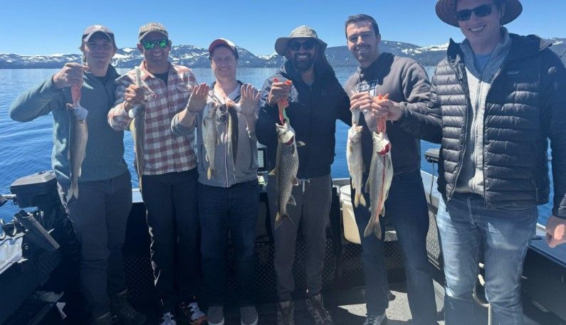 Six people on a boat display their caught fish on a lake under a sunny sky.