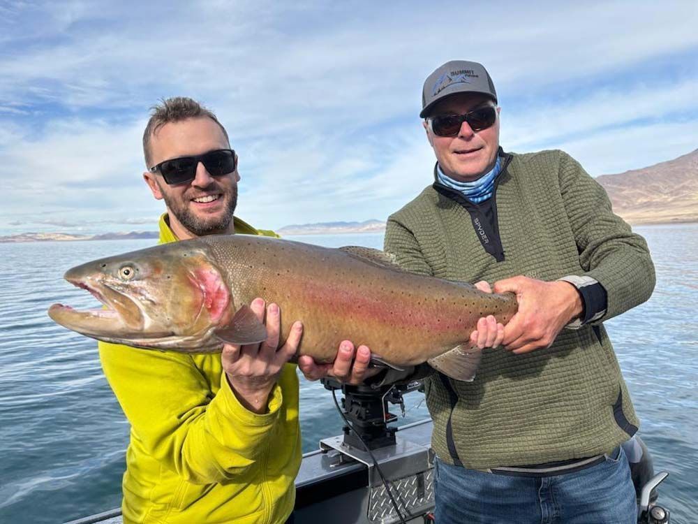 Two men on a boat hold up a large, colorful trout caught in a lake.