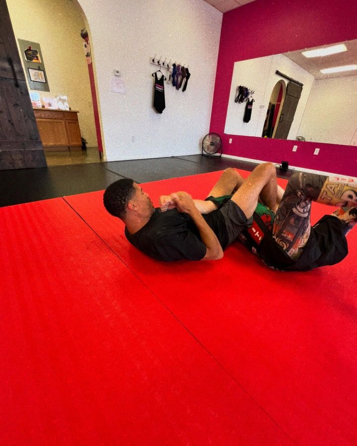 Two people grappling on a red mat in a martial arts studio with white walls and mirrors.