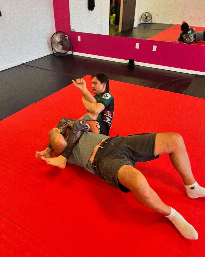 Two people training on red mats in a martial arts gym, one lying back while the other reaches forward.