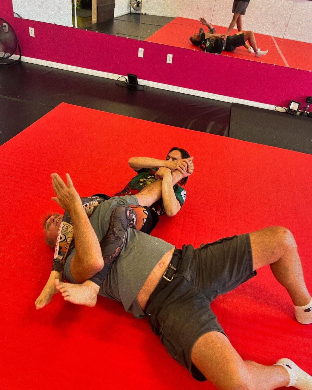 Two people wrestling on red mats in a gym, one pinned on the floor with a mirrored wall behind them