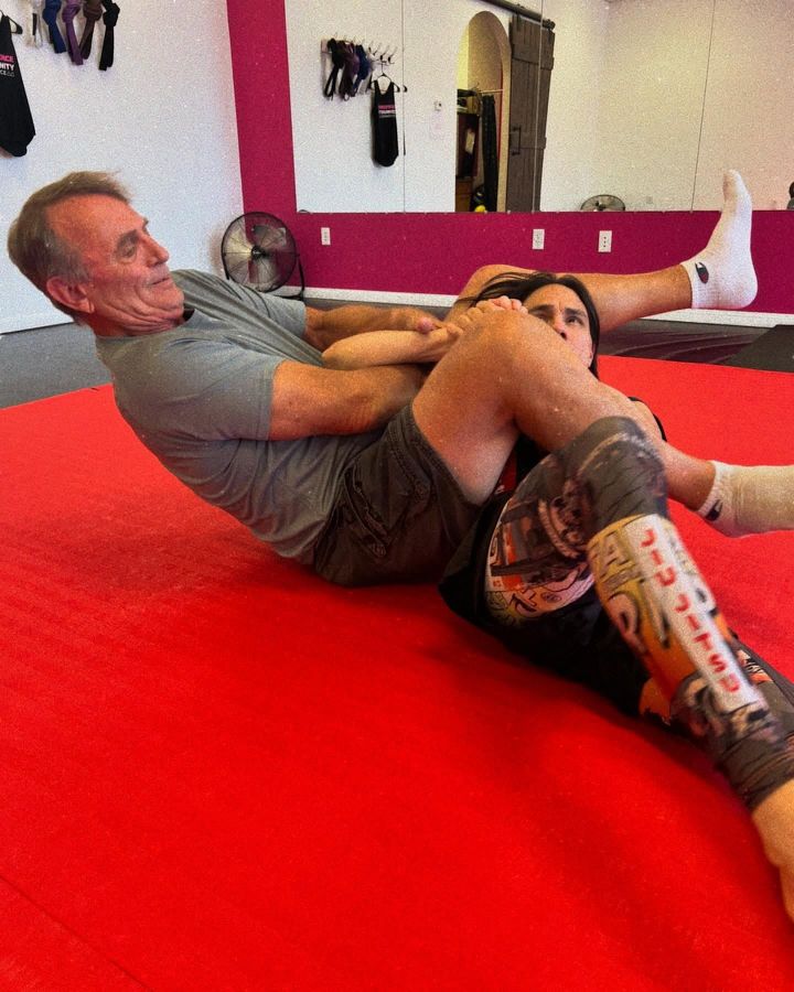 Two people grappling on red mats in a martial arts gym, one holding a leg lock.