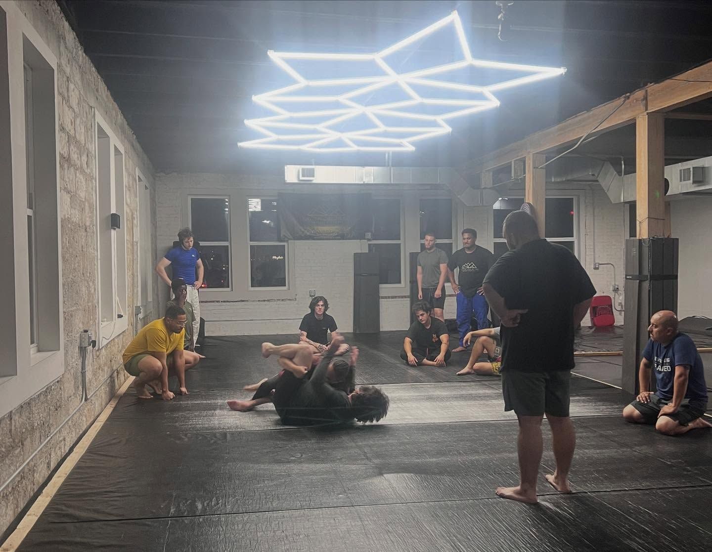 People training in a martial arts gym on mats under bright geometric ceiling lights.