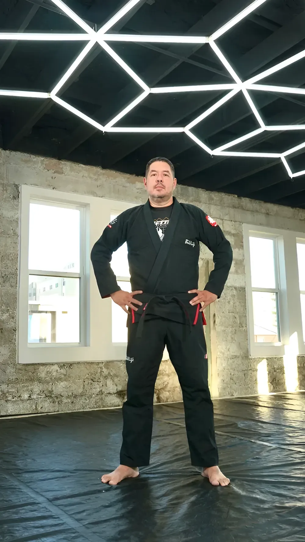 Person standing barefoot in a black martial arts uniform in a bright studio with geometric ceiling lights.