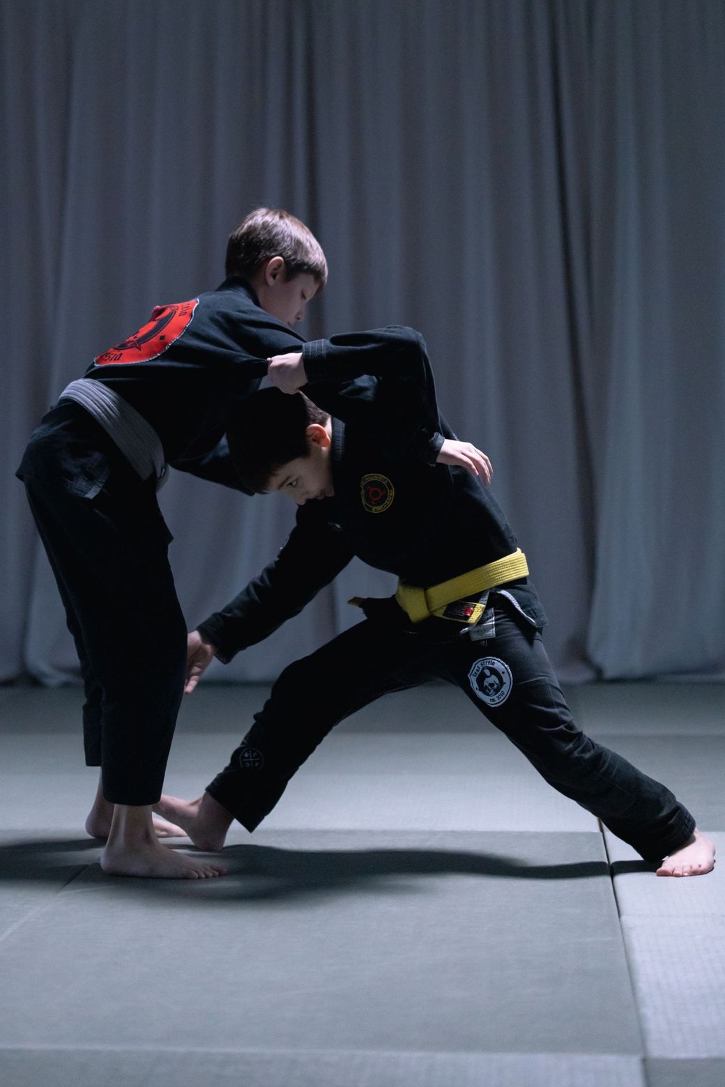 Two martial artists sparring on a mat, one in a black gi with a yellow belt, against a gray curtain backdrop