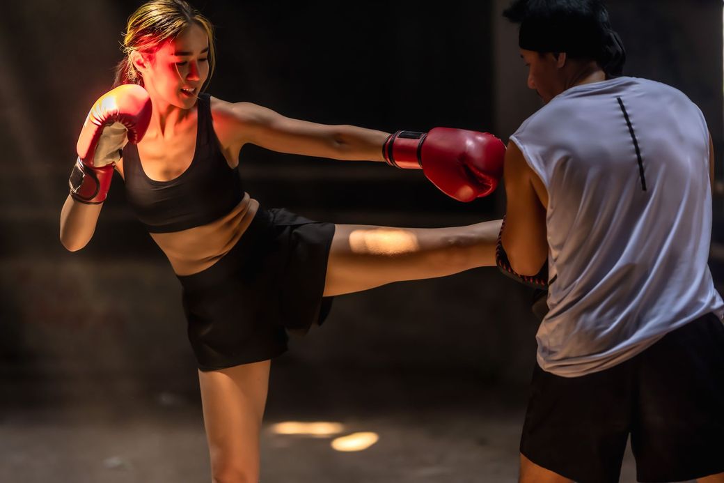 Two kickboxers sparring, woman in red gloves landing a high kick on an opponent in a white shirt