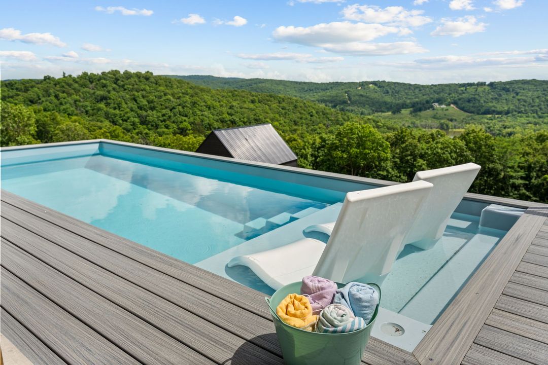 Poolside lounge chair on a wooden deck beside calm water, with a large umbrella and grassy lawn behind.