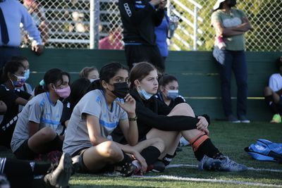 Soccer players in masks sit on the field, watching the game.