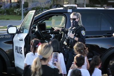 Police officer in uniform points at equipment in patrol car, children and adult look on.