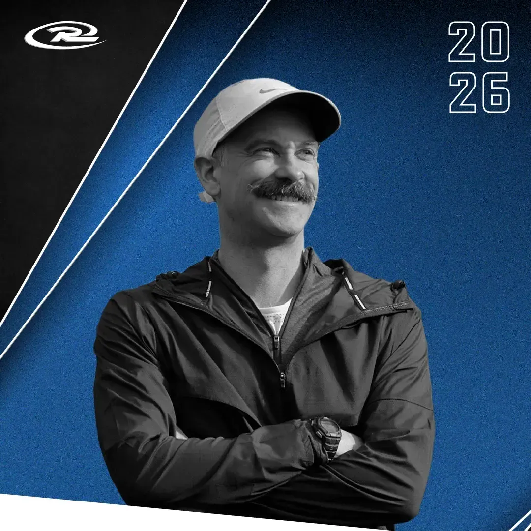 Smiling man in a cap and jacket with arms crossed on a blue 2026 background
