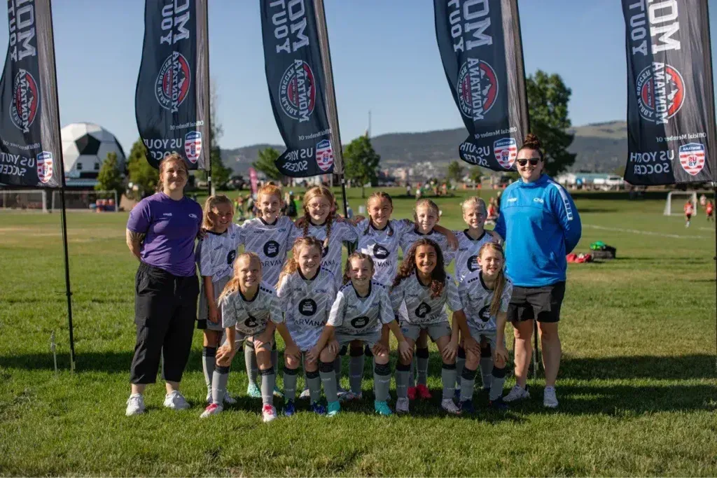 Youth soccer team poses on a grassy field with coaches and flags; sunny day.