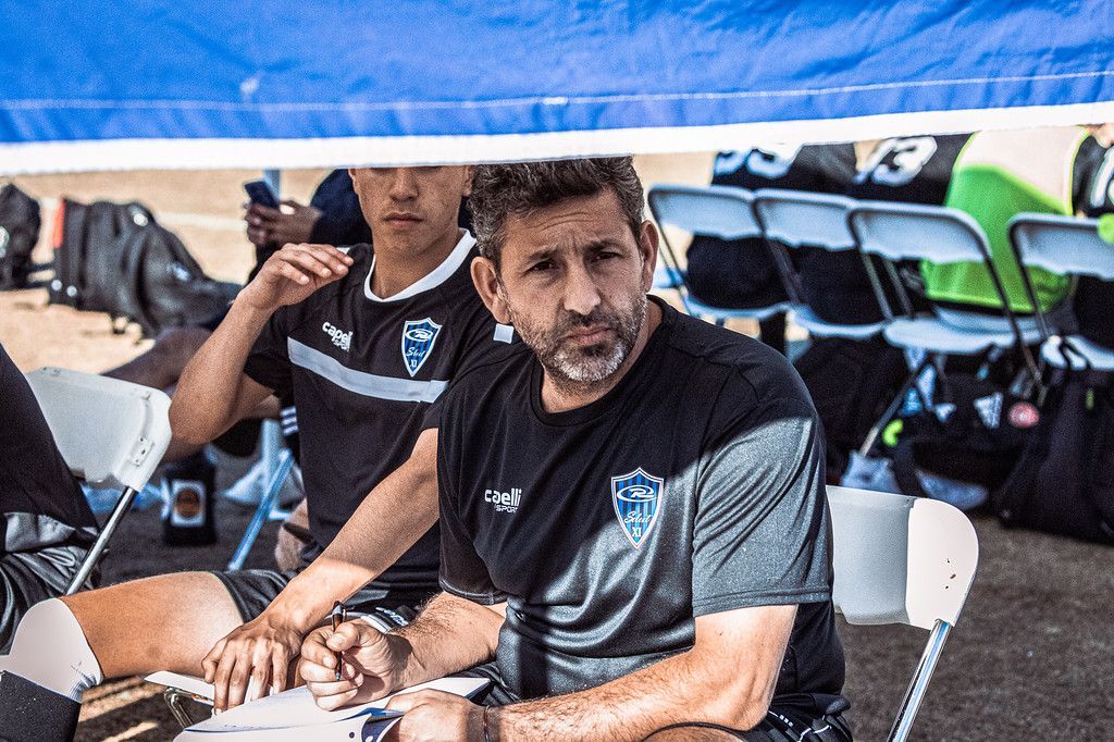 Soccer coach and player seated under a canopy, reviewing notes during a game, team in background.