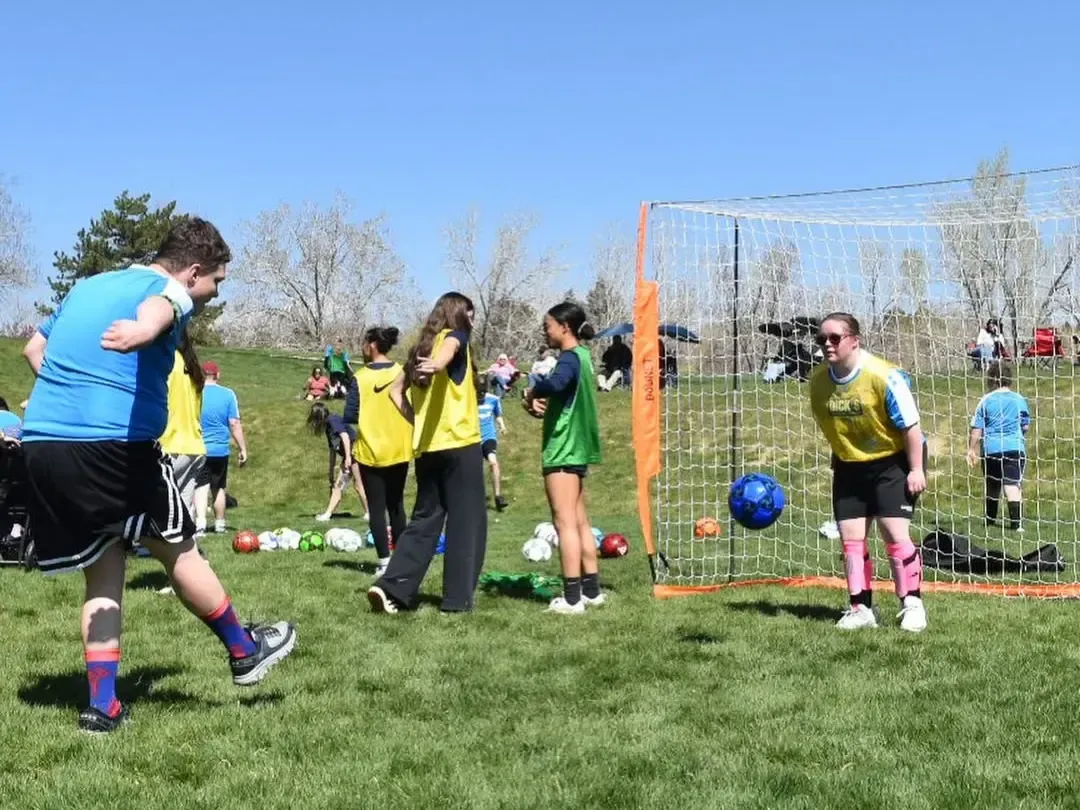 Soccer players on a grassy field; one kicking a ball towards a net; others watching. Sunny day.