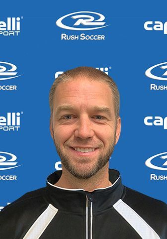 Man with short hair and beard smiles in front of a blue backdrop with soccer logos.