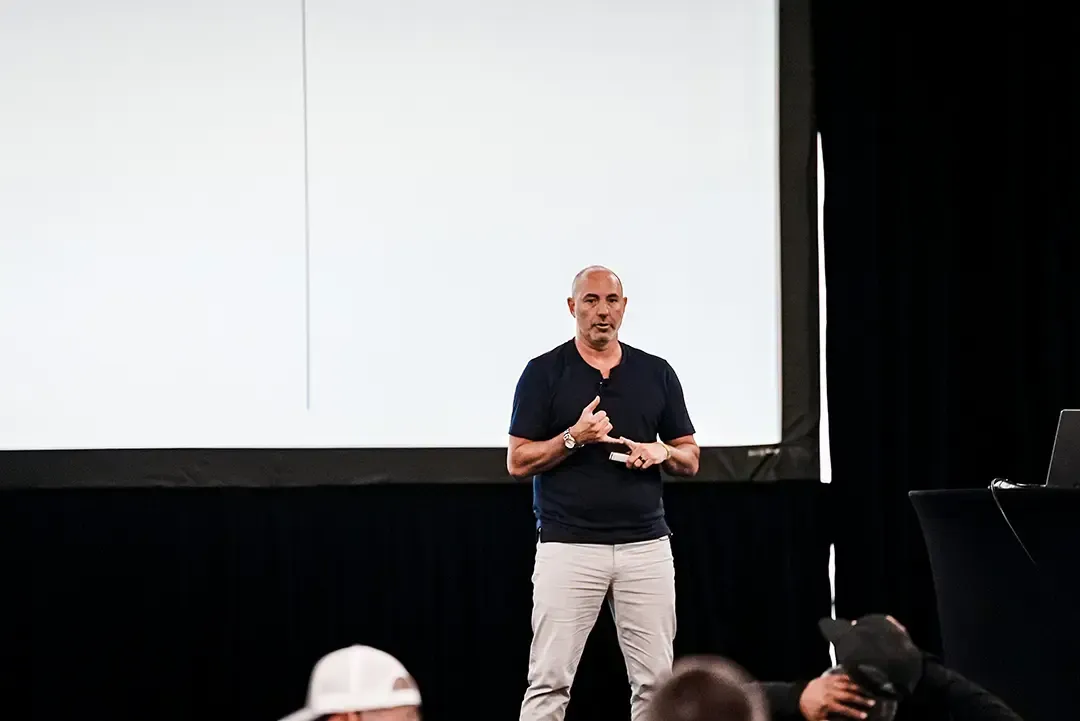 Man speaking on a stage in front of a large blank screen, with audience heads visible in foreground.