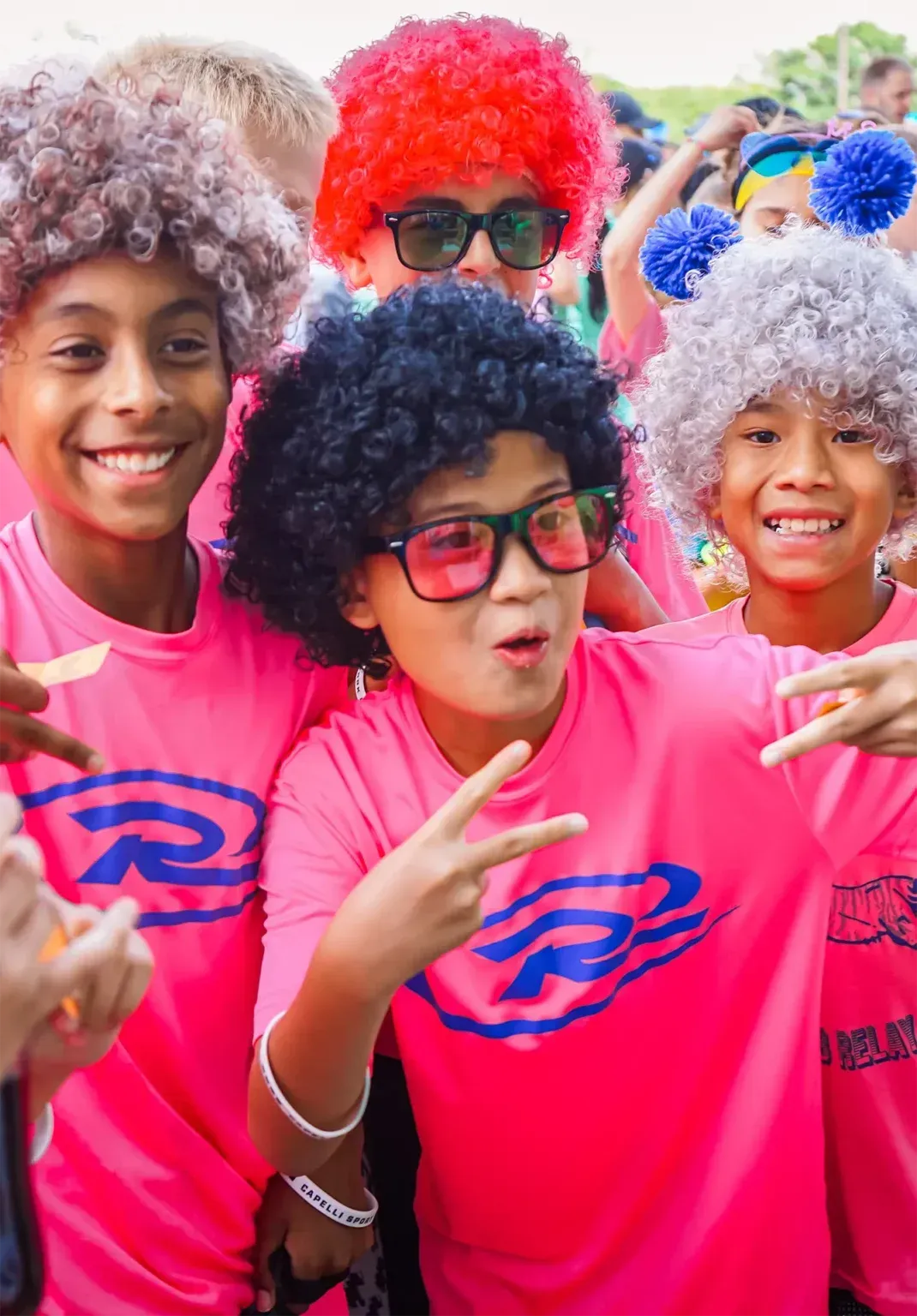 Children in pink shirts and afro wigs smiling, making peace signs, outdoors.