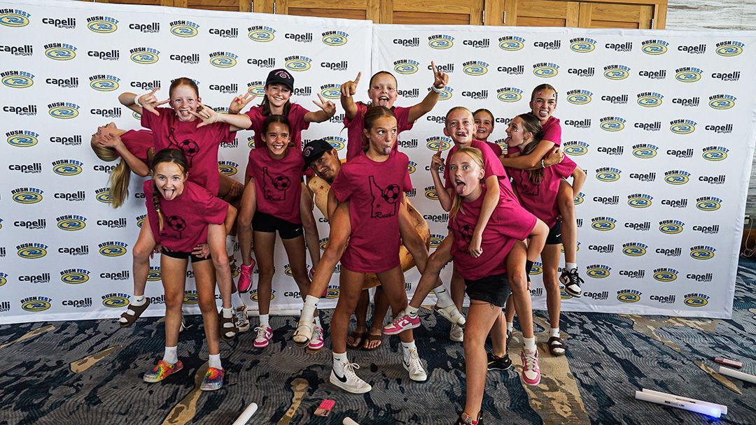 A group of people in burgundy shirts and shorts pose excitedly in front of a backdrop, some giving piggyback rides.