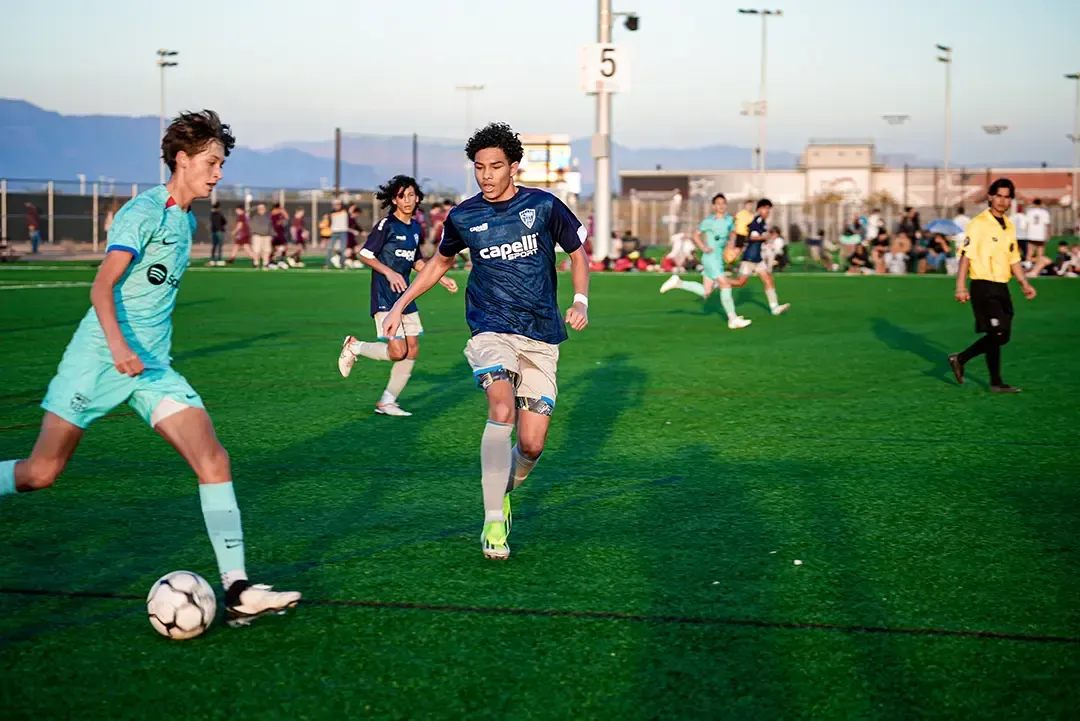 Soccer game on a green field; players in blue and light blue uniforms, running and kicking the ball.