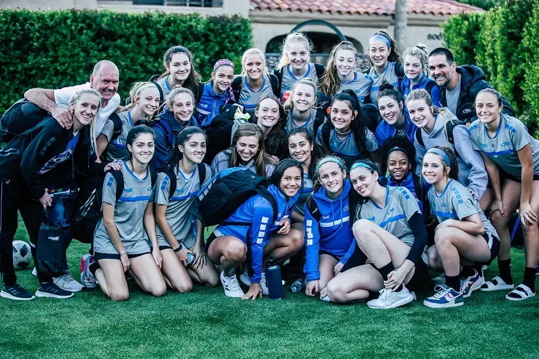 Soccer team posing together on a grassy lawn. Group smiles at the camera. Two coaches are present.