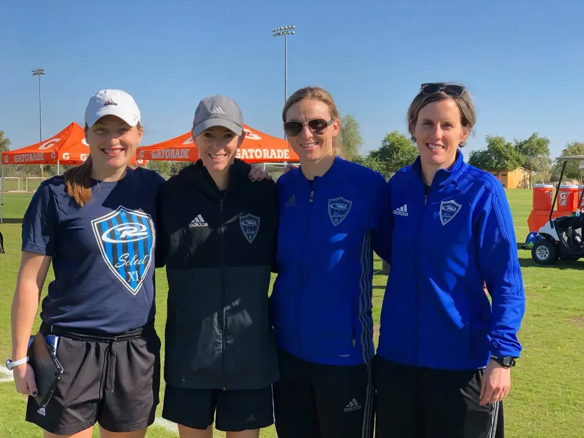 Four people in sports attire pose outdoors. Two wear blue, one a referee uniform, and one a cap.