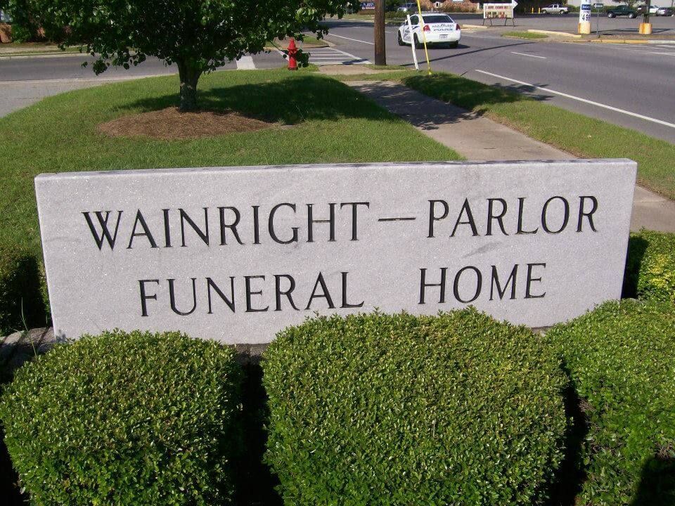 Stone sign reading “Wainright-Parlor Funeral Home” beside trimmed hedges and a roadside.