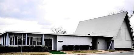 Single-story white church building with a steep roof and dark windows, seen from the front.
