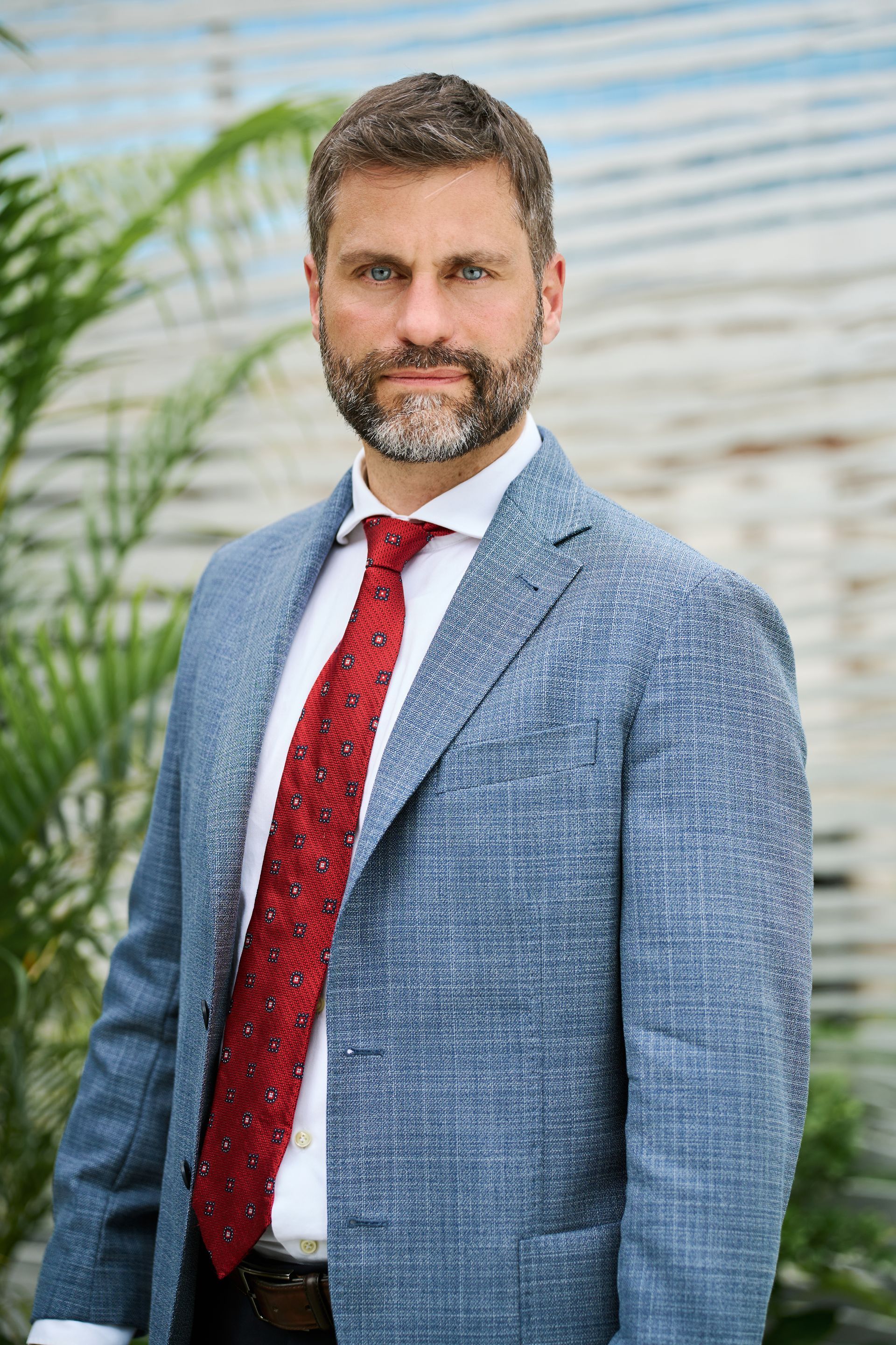 Man in blue blazer, red tie, standing outdoors near green plants, looking at the viewer.