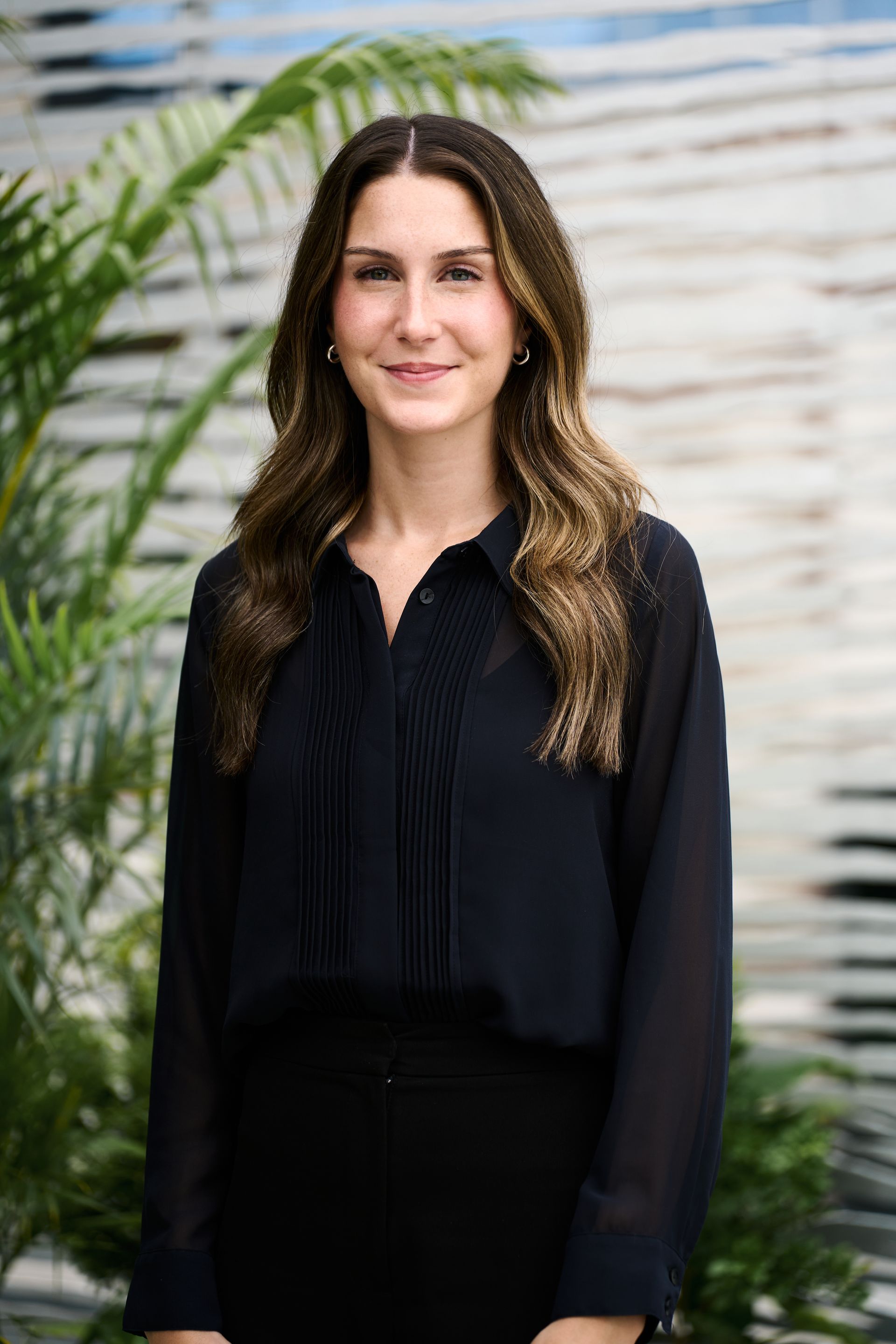 Woman with long brown hair in a black shirt and skirt, smiling. Standing outside, greenery in background.