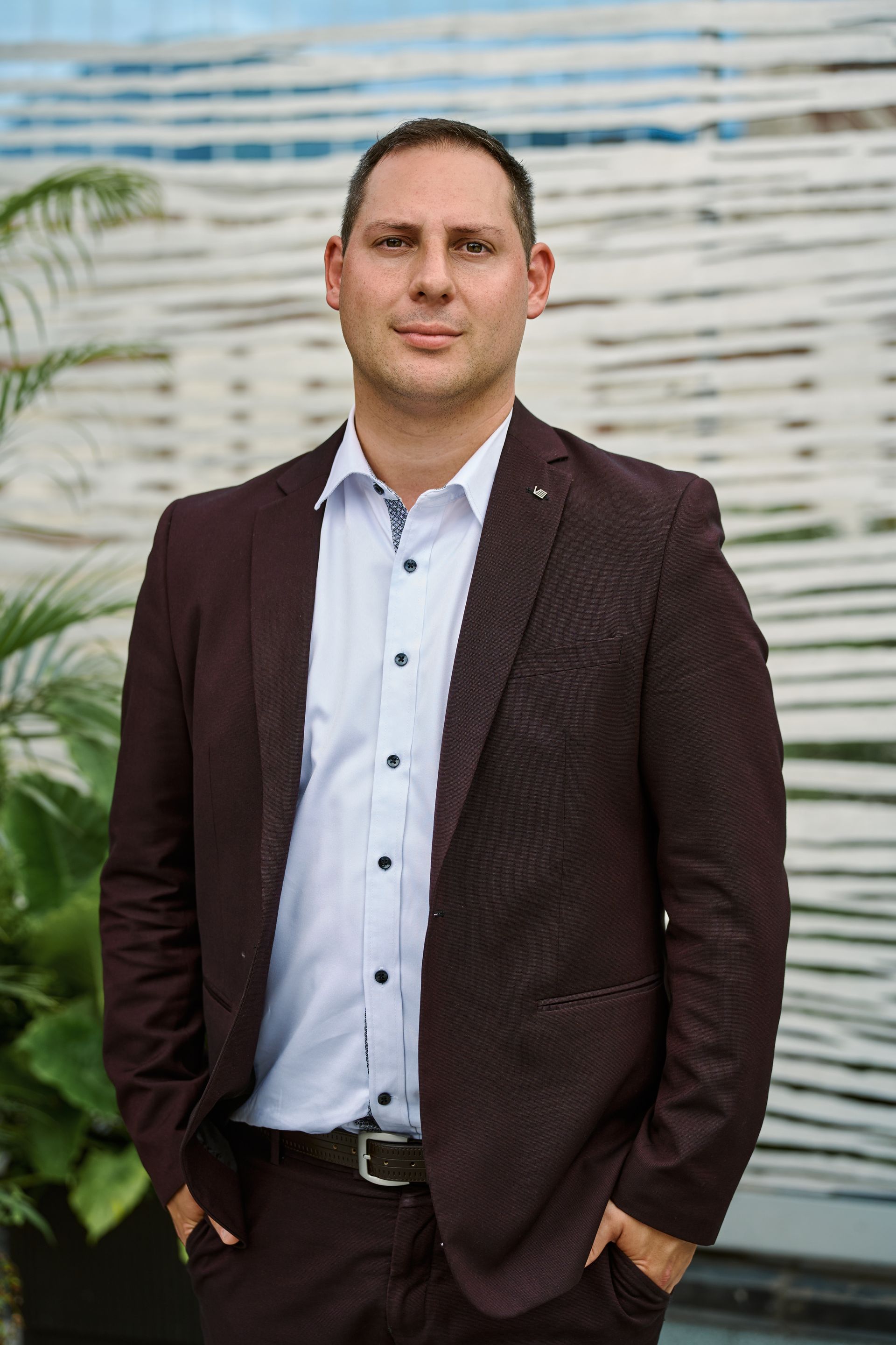 Man in maroon suit and white shirt, hands in pockets, smiling. Outdoor setting.