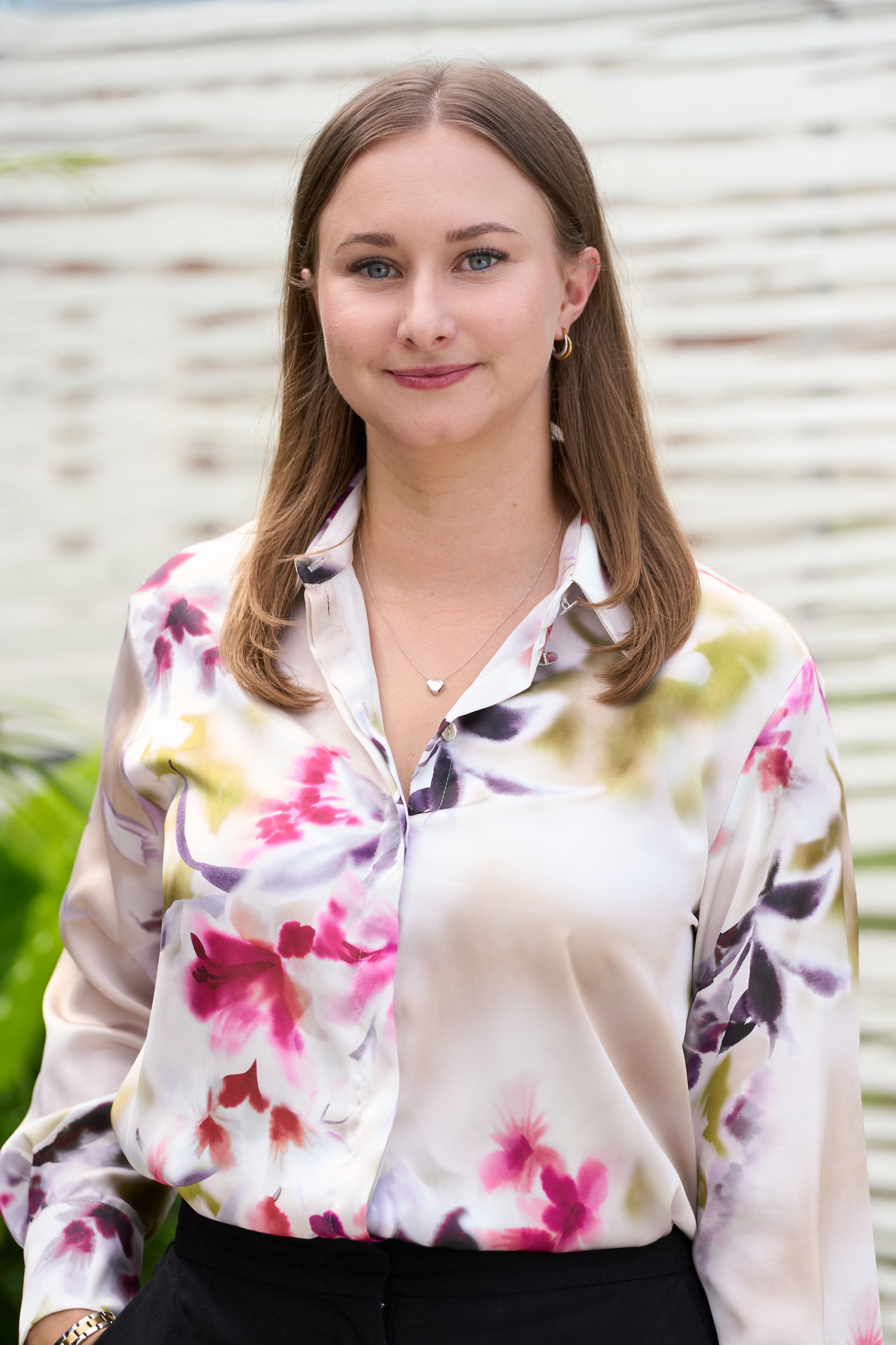 Young woman with blonde hair, wearing a floral blouse, smiling, against a blurred background.