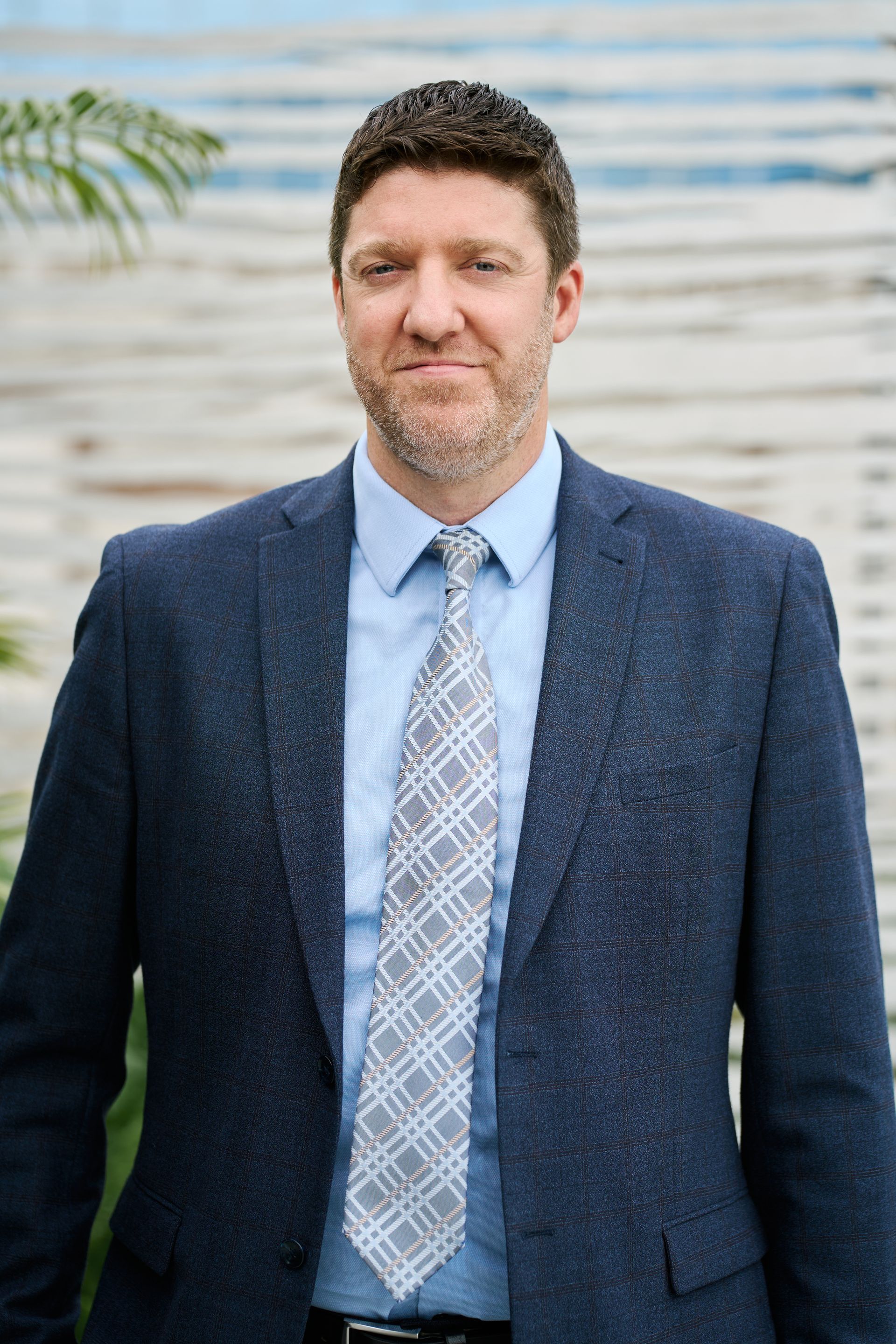 Man in blue blazer and patterned tie smiles, standing in front of a light wooden wall.