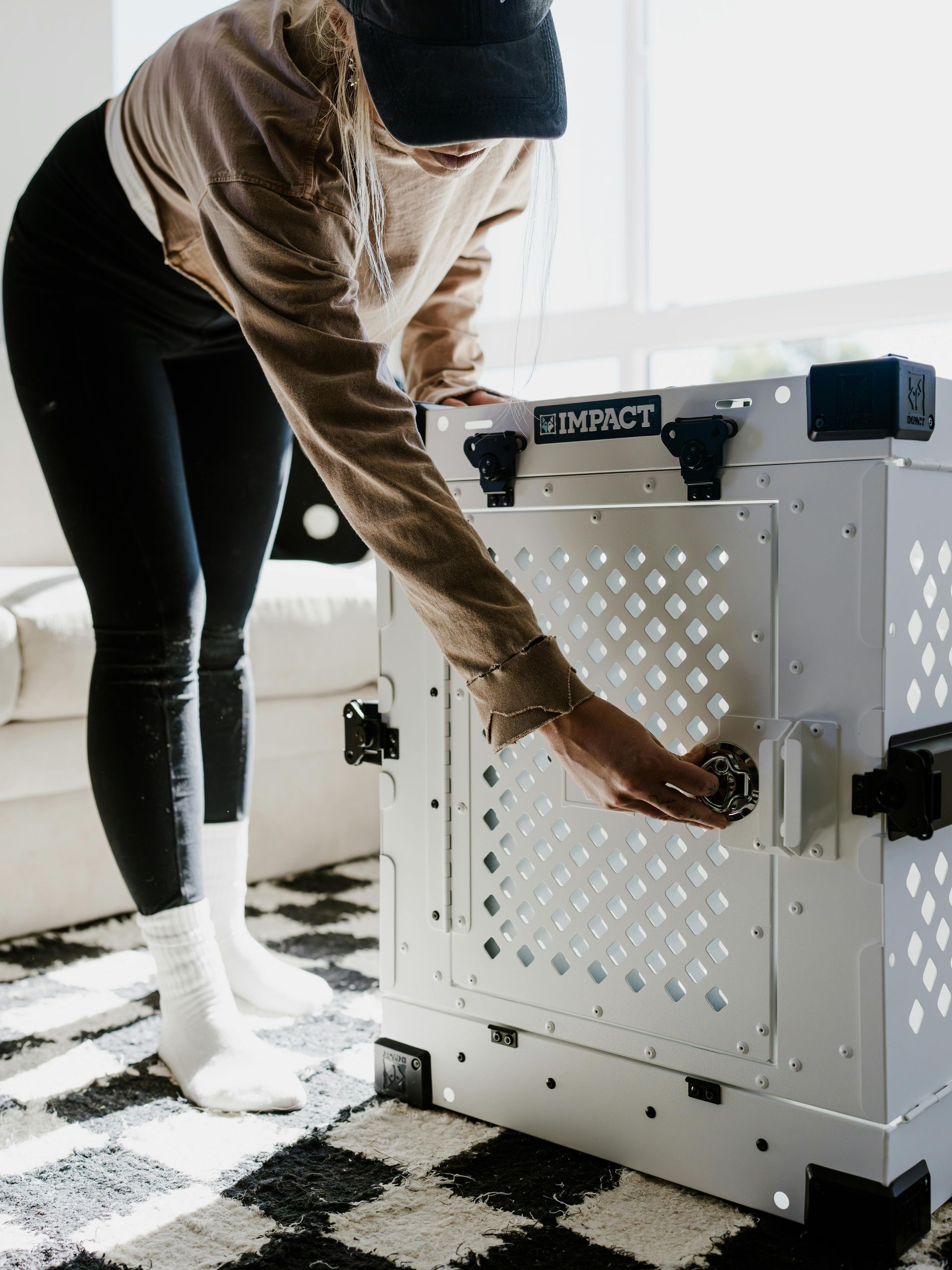 Een persoon in een beige top en zwarte legging opent de deur van een witte metalen hondenbench van het merk Impact, die op een geruite mat staat.