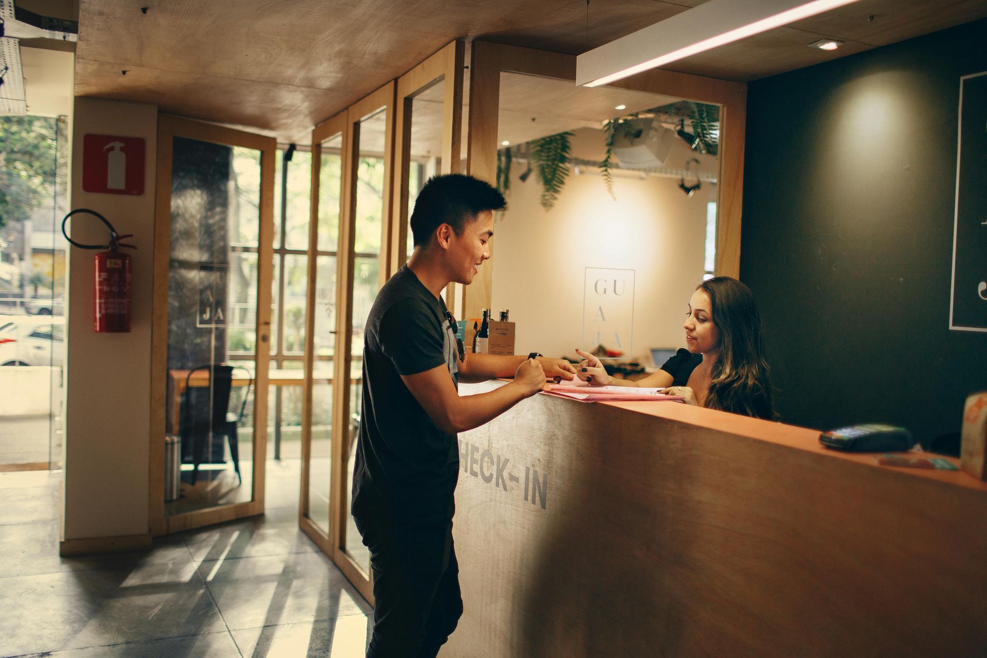 Een persoon staat bij een houten receptiebalie in gesprek met een receptioniste in een lichte, moderne ruimte.
