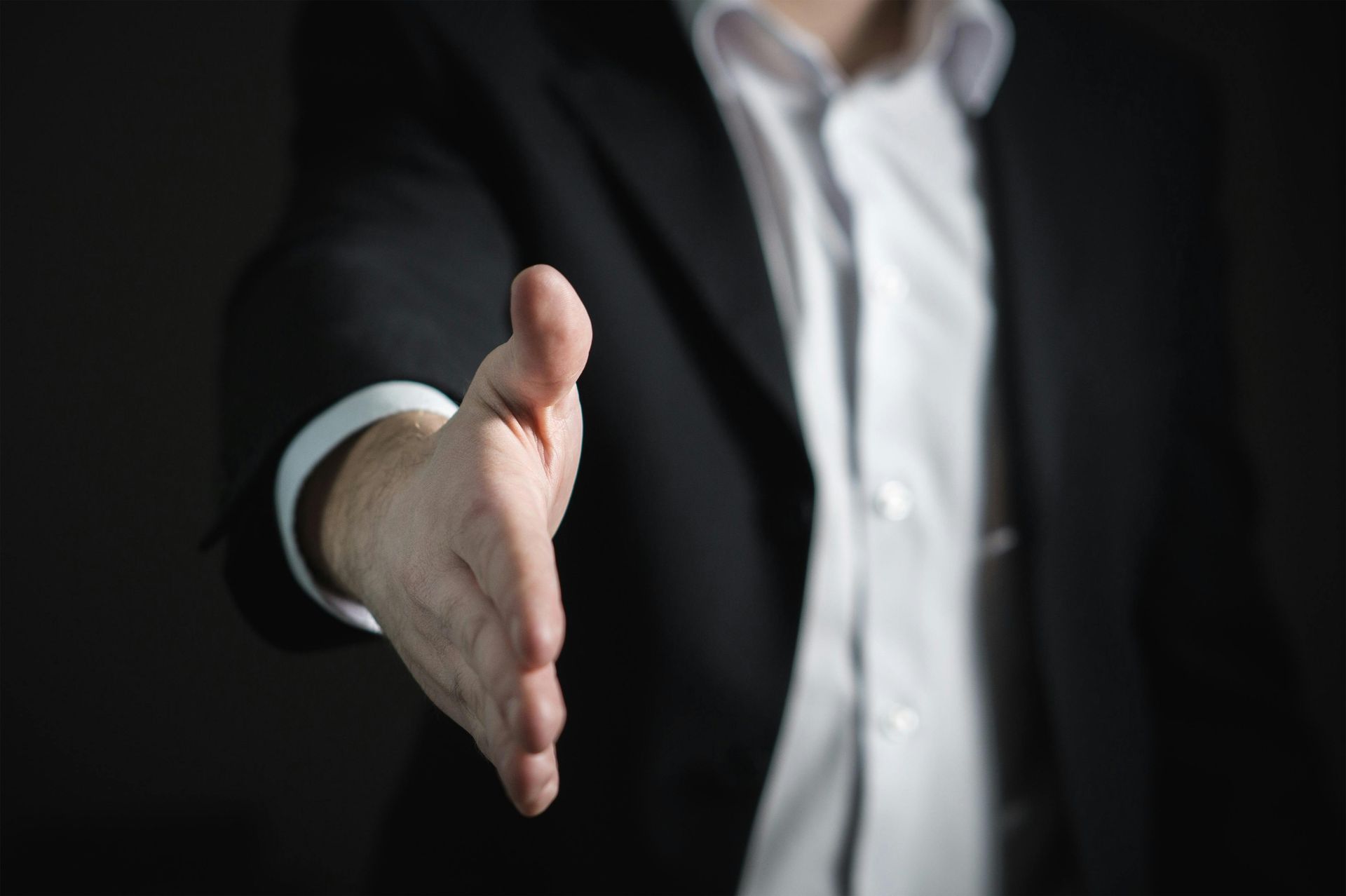 Een persoon in een businesspak steekt zijn hand uit voor een handdruk tegen een donkere achtergrond.