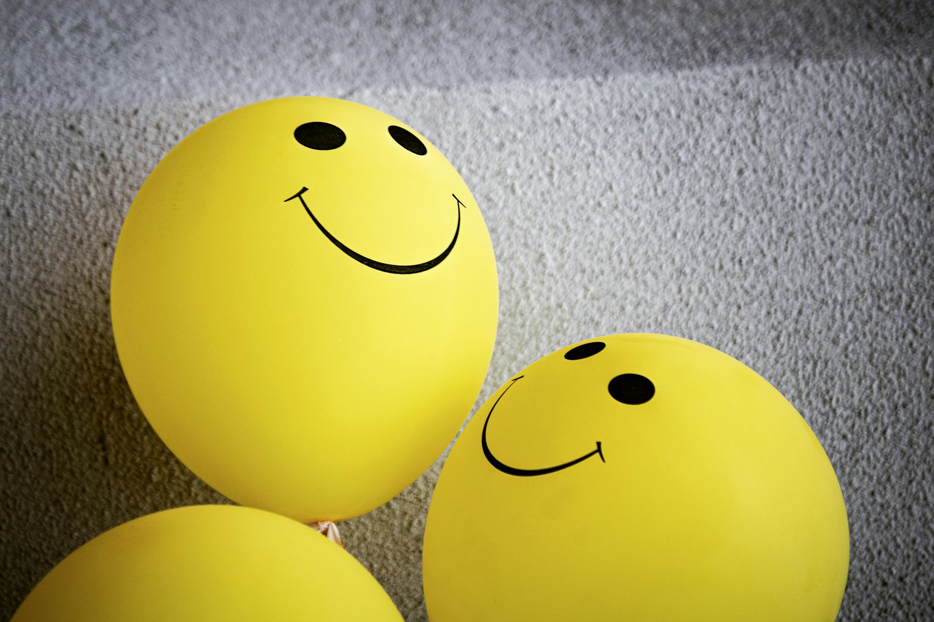 Drie gele, ronde ballonnen met zwarte smileygezichtjes staan bij elkaar tegen een grijze, gestructureerde achtergrond.