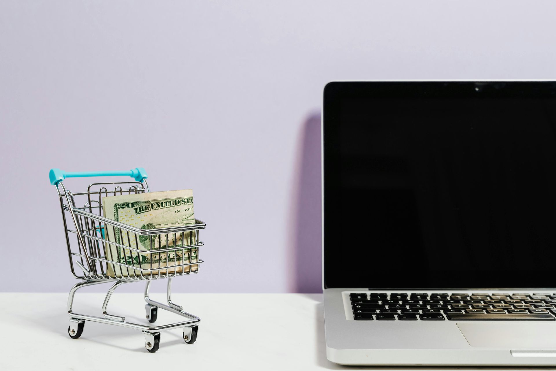 Een miniatuurwinkelwagentje gevuld met Amerikaanse dollarbiljetten staat op een wit oppervlak naast een opengeklapte laptop.