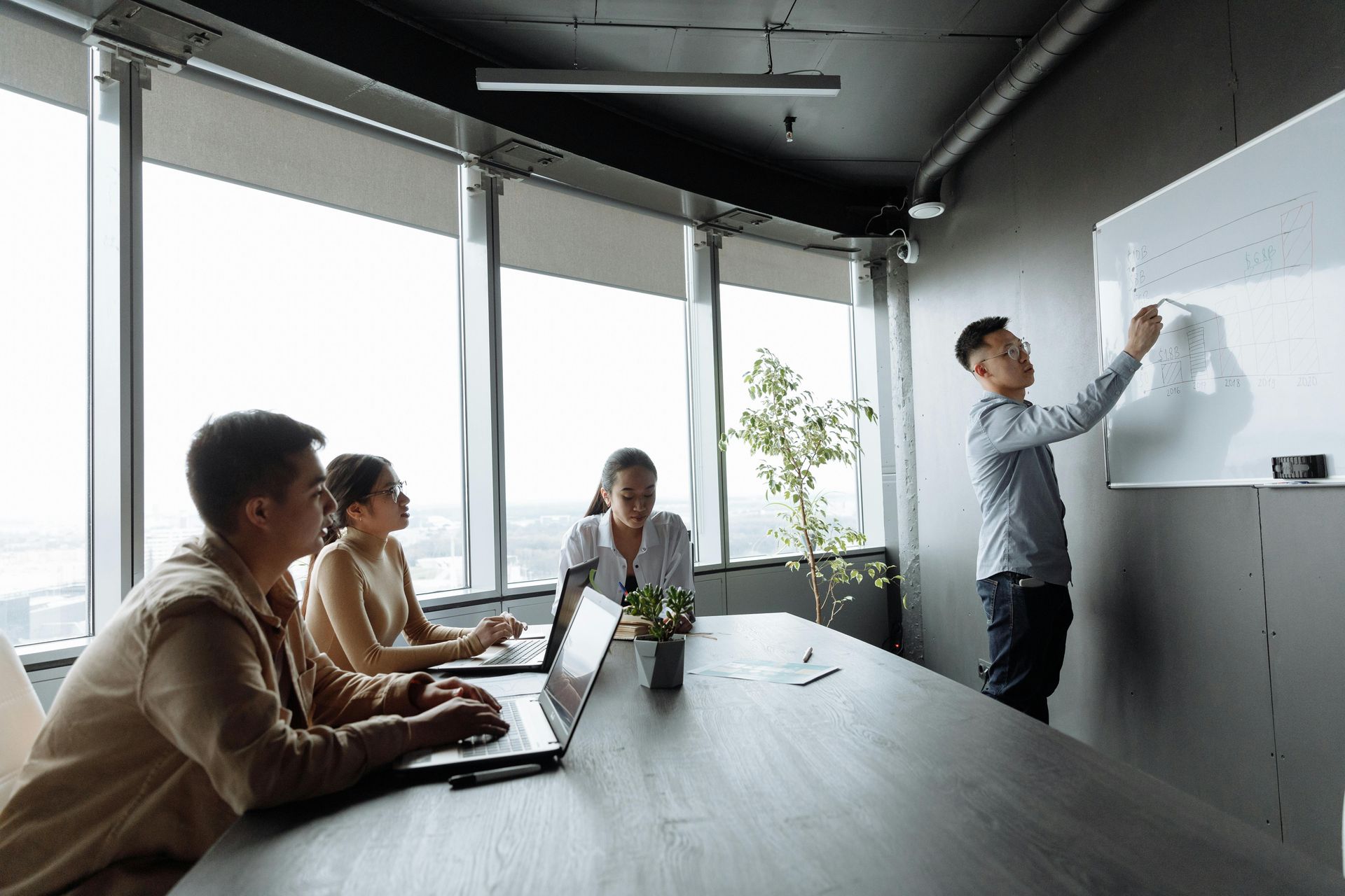Tijdens een vergadering met drie collega's die met laptops aan een tafel zitten in een licht kantoor, schrijft iemand op een whiteboard.