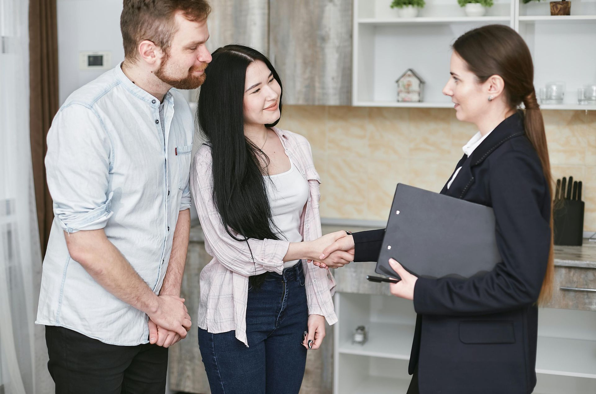 Een makelaar in een donkere blazer schudt iemand de hand, terwijl een partner toekijkt in een felverlichte keuken.