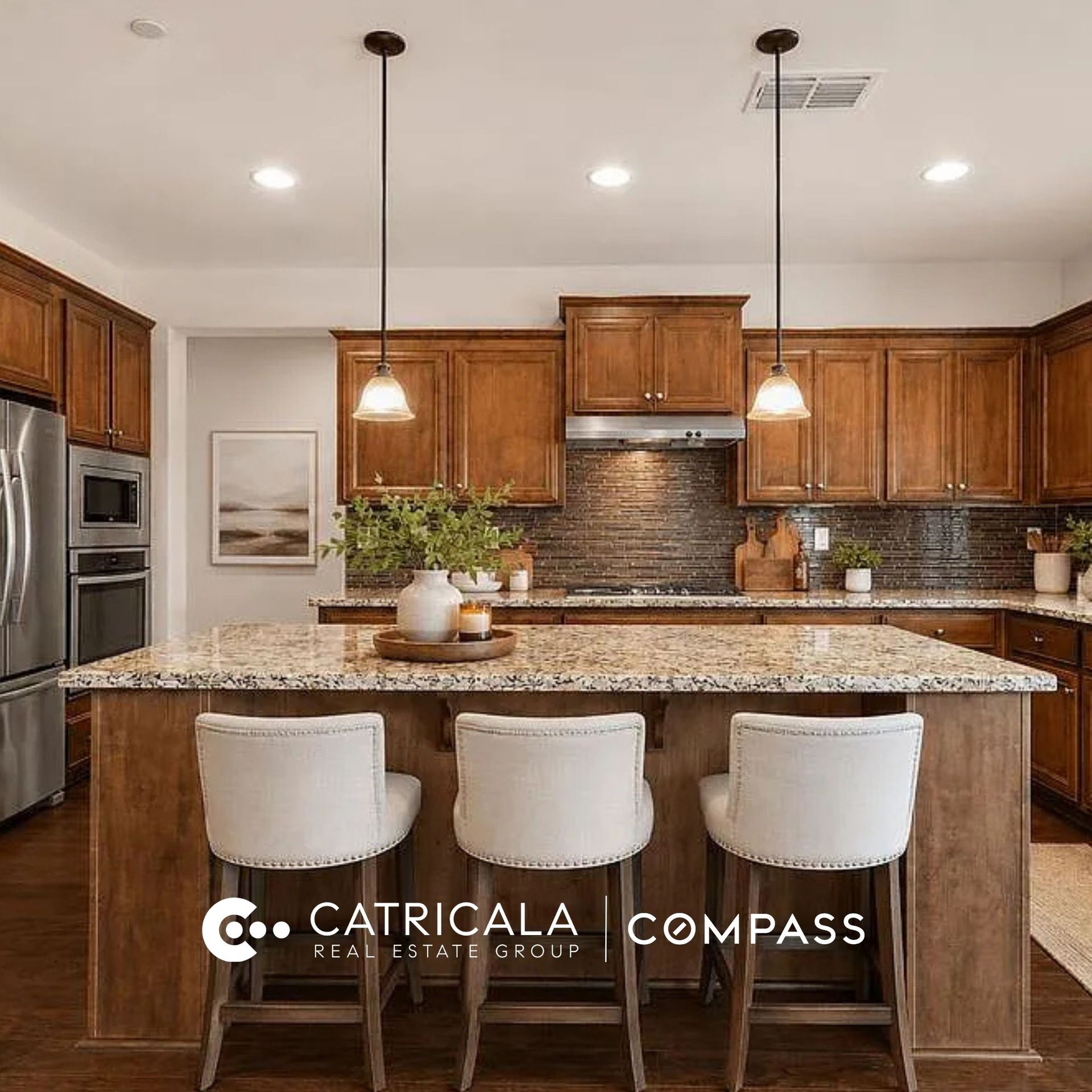 Kitchen featuring wood cabinetry, a granite island with three white stools, and the Catricala Real Estate Group Compass logo.