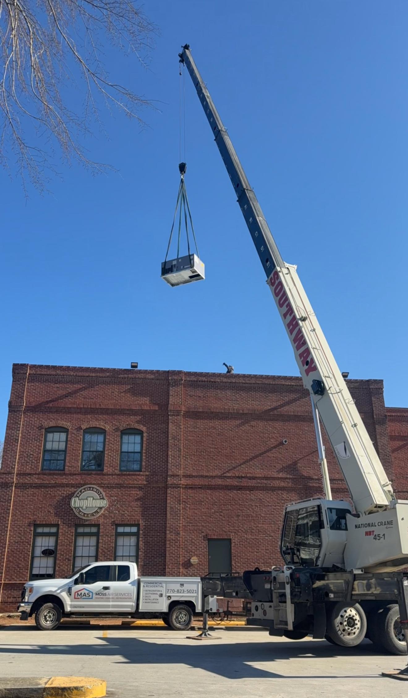 Crane lifting a white item above a brick building beside a parked pickup truck under a blue sky