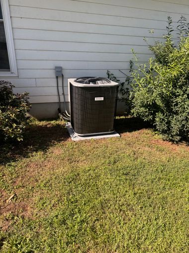 Outdoor air conditioner unit beside a white house wall, partly surrounded by grass and bushes.
