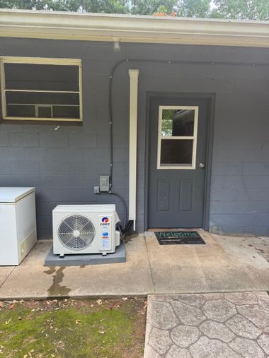 Gray house exterior with a window, door, outdoor AC unit, and small doormat on a concrete porch.