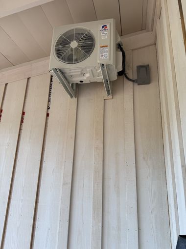 Wall-mounted outdoor air conditioner unit with a fan on a beige wooden wall under a roof