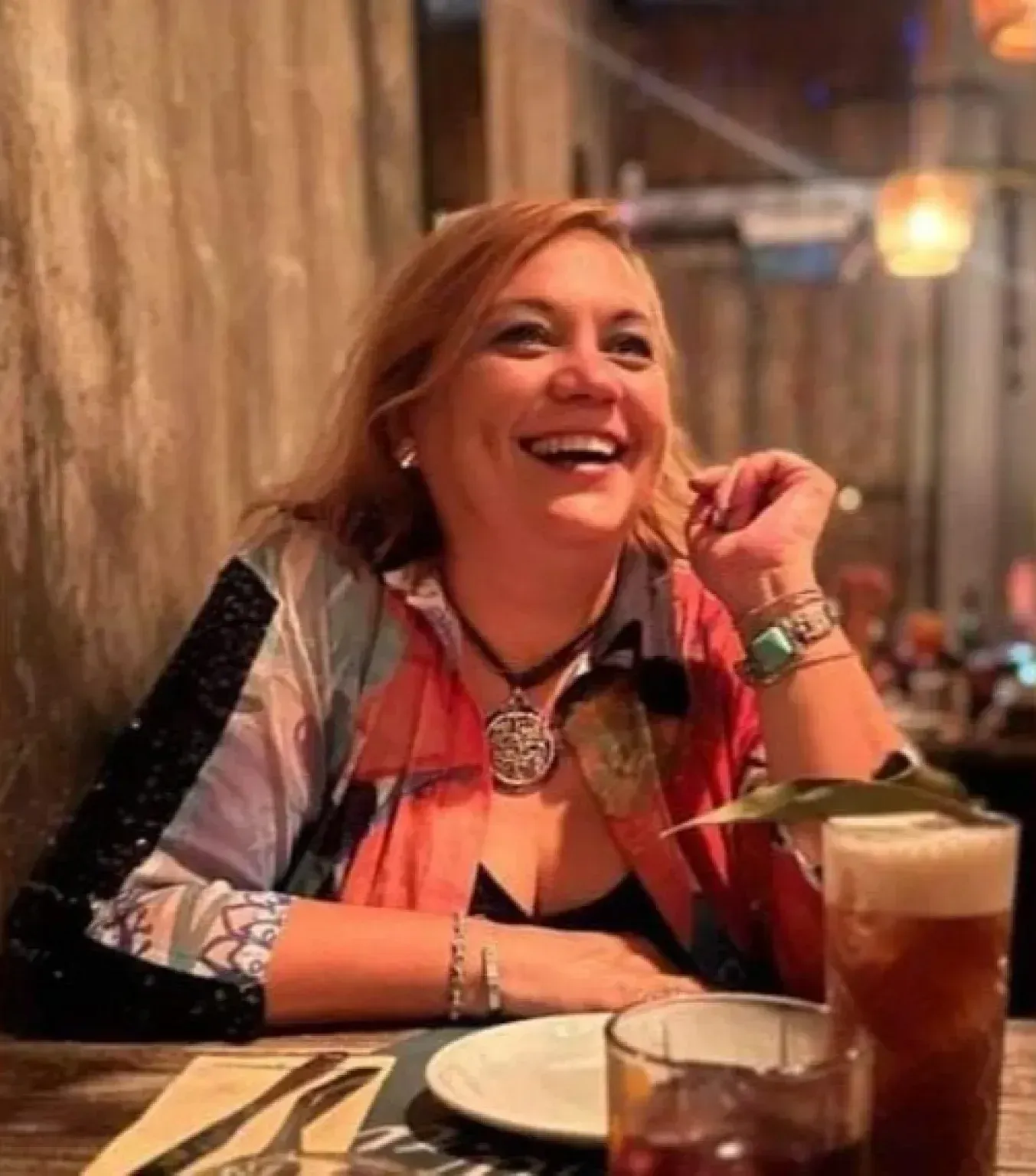 Una persona sonriente sentada en la mesa de un restaurante con una bebida, vistiendo una blusa colorida y un collar.