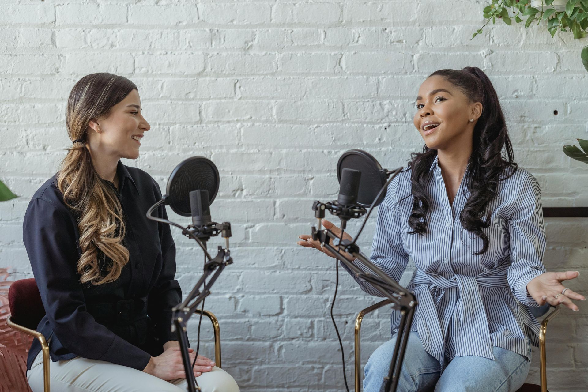 Two women recording a podcast in front of a white brick wall. They are speaking into microphones.