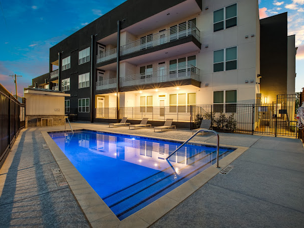 Angular shot of real estate property with a blue lit pool. The sun is setting behind the property with a dark grey and white facade. 