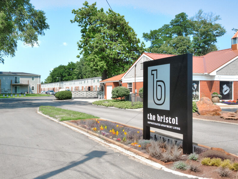 Wide shot of the entrance to a residential complex with one-story brick and sided buildings. Green tree surrounding the entry.