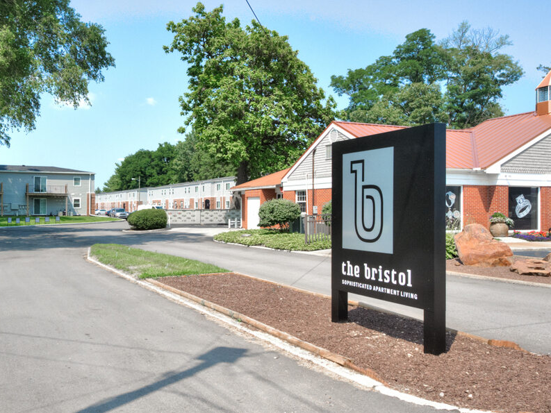 Wide shot of the entrance to a residential complex with one-story brick and sided buildings in Indiana. Green trees surrounding the drive-in entry.