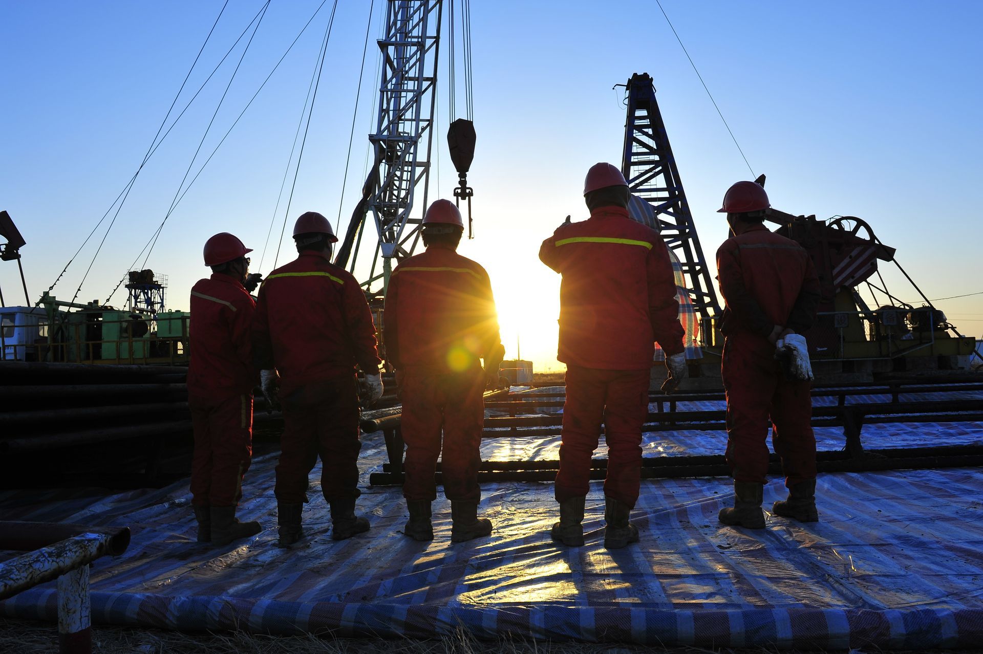 Five workers in red coveralls and hard hats stand before an oil drilling rig silhouetted against a bright sunrise.
