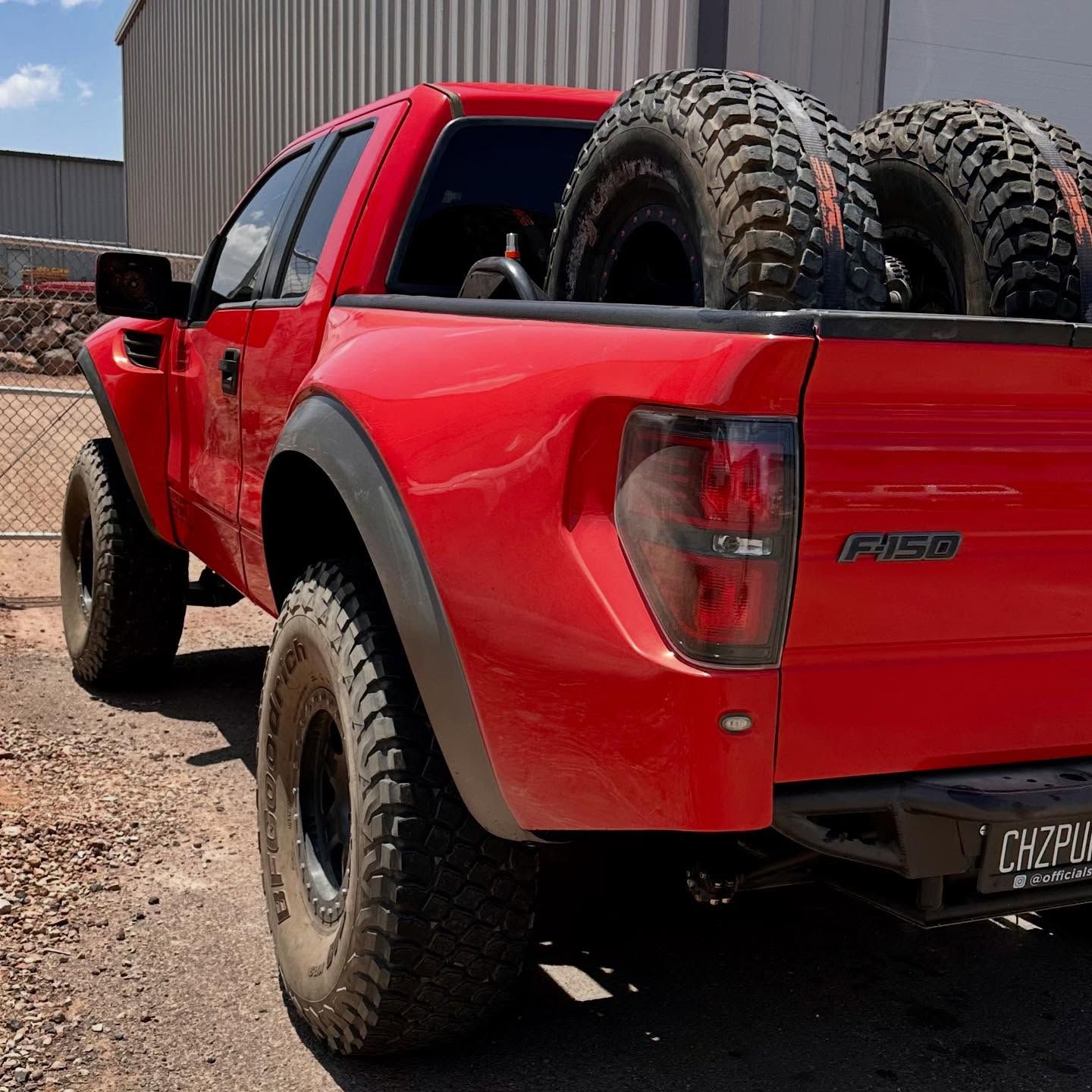 A red Ford F-150 off-road truck parked outside, with two large spare tires secured in its bed.